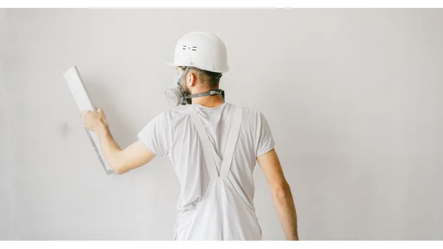 Back view of a construction worker in safety gear plastering a white wall.