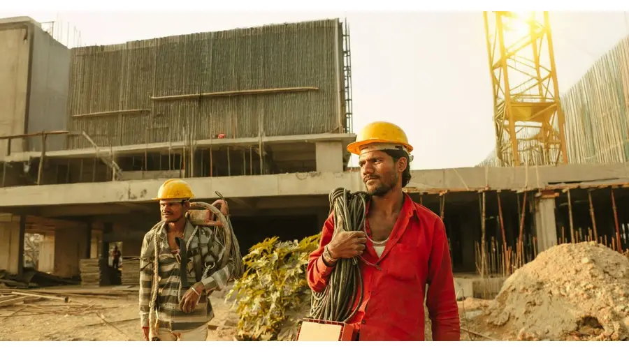 Two construction workers wearing helmets at a site in India with a crane and building.