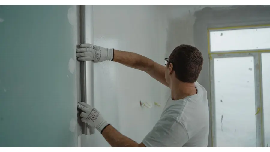 A man in a white shirt and gloves using a metal ruler for indoor wall renovation work.